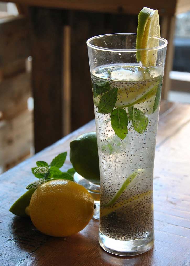 vaso alto con limonada de chía con hojas de menta y dos limones en la mesa